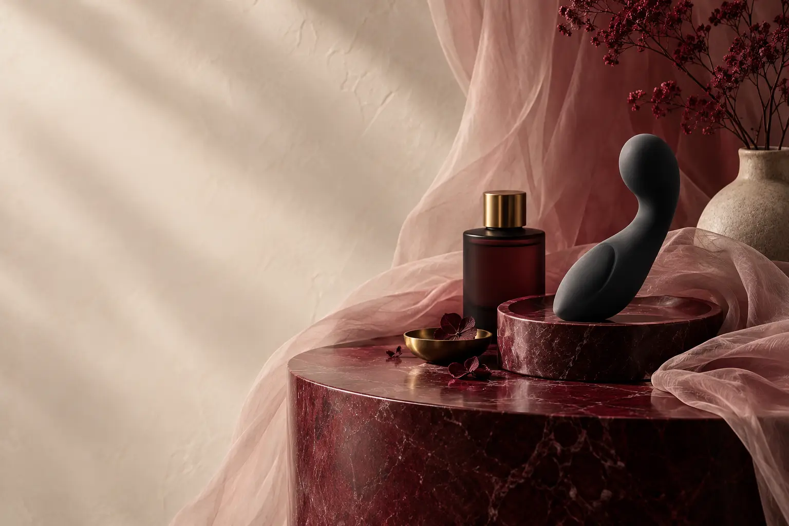 Illustrative still life: a dark matte object on a burgundy marble plinth beside a dark glass bottle, pink tulle and dried flowers